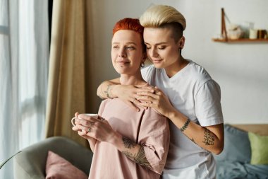 Kısa saçlı iki kadın, LGBT yaşam tarzındaki aşkın güzelliğini yansıtan rahat bir oturma odasında birbirlerine sarılıyorlar..