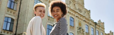 A stylish multicultural lesbian couple stands gracefully in front of a university building. clipart