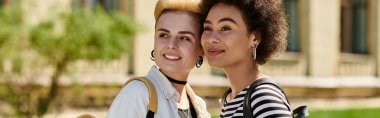 Çok kültürlü, şık giyinmiş lezbiyen bir çift üniversite kampüsündeki bir binanın önünde duruyor..