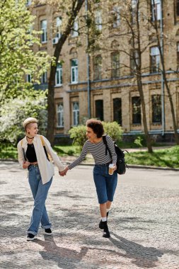 İki genç kadın, el ele tutuşarak, bir üniversite kampüsünün yakınındaki kaldırım taşı caddesinde yürüyorlar..