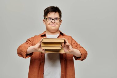 Down sendromlu bir çocuk mutlu bir şekilde gri arka planda bir yığın kitap tutuyor..