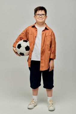 Down sendromlu gözlüklü küçük bir çocuk futbol topu tutuyor..