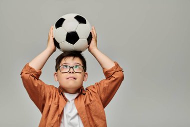 Down sendromlu küçük sevimli bir çocuk neşeyle başının üstünde bir futbol topu tutuyor..