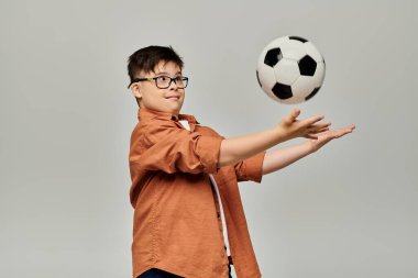 Down sendromlu çekici bir çocuk gri arka planda bir futbol topunu dengeliyor..