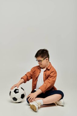 Down sendromlu küçük çocuk elinde futbol topuyla yerde oturuyor..