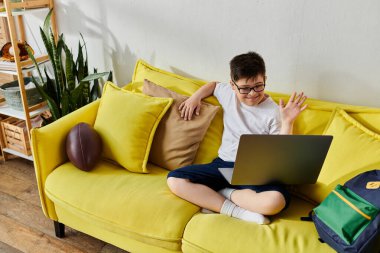 adorable boy with Down syndrome uses laptop on yellow couch at home. clipart