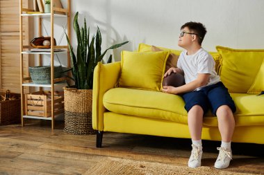 Down sendromlu sevimli bir çocuk odasında canlı sarı bir kanepede mutlu bir şekilde oturuyor..