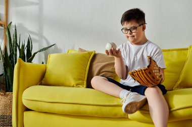 Down sendromlu çekici bir çocuk elinde beyzbol eldiveniyle güneşli sarı bir kanepede yatıyor..