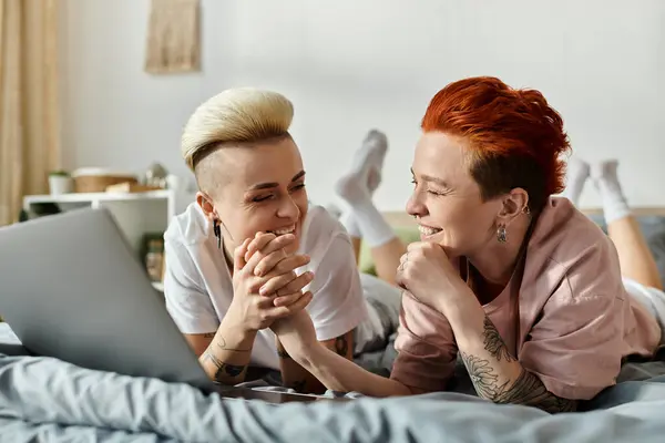 stock image Two people, a lesbian couple with short hair, are laying on a bed together and looking at a laptop screen in a cozy bedroom setting.