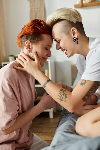 A lesbian couple with short hair sitting on a bed, exchanging smiles and radiating joy in their intimate bedroom setting.