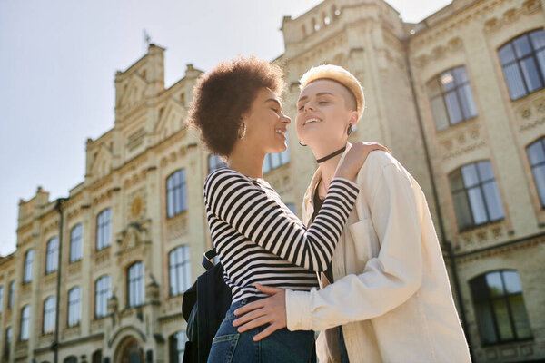Two young women dressed stylishly embrace in front of a stunning urban building, showcasing their close connection.