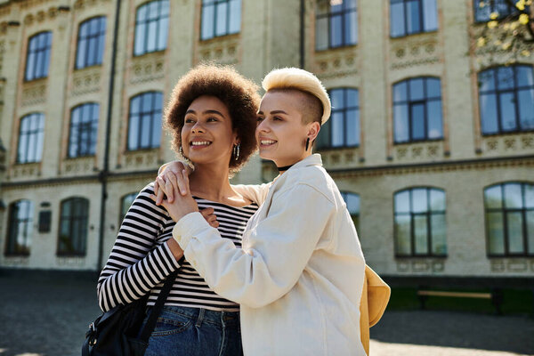 Two young women in casual attire embrace in front of a university building.