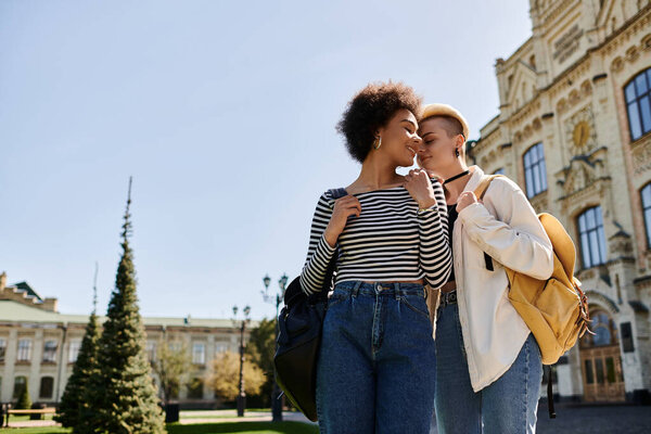 A stylish multicultural lesbian couple, in casual attire, stands in front of a building on a university campus.