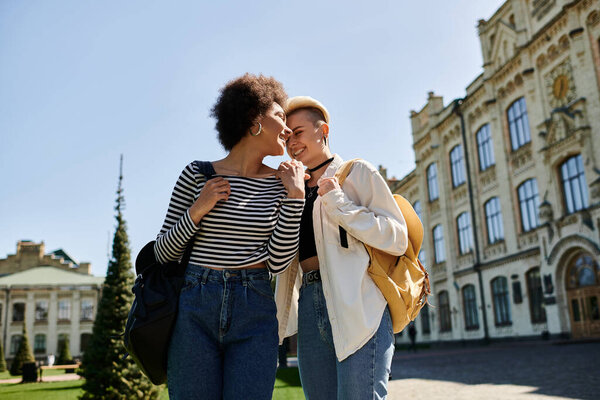 Two young women in fashionable attire share a kiss in front of a bustling building on a university campus.
