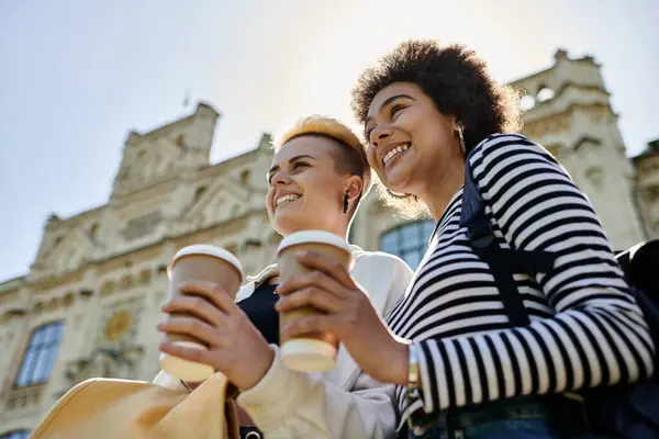 stock image Two young women, casually dressed, sipping coffee in front of a majestic building.