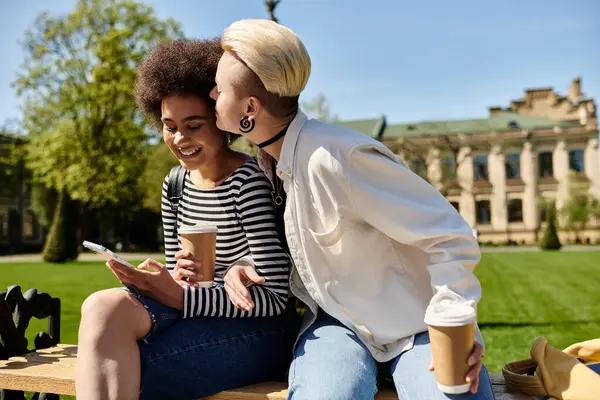 stock image Two young women in stylish attire are seated on a park bench, deep in conversation, surrounded by greenery.