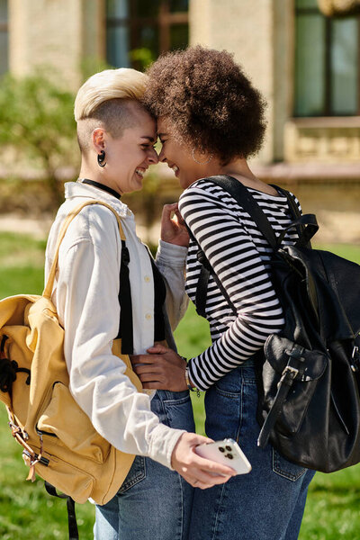 Two young women with backpacks sharing a joyful moment, smiling at each other outdoors near a university campus.