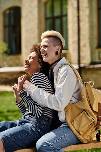 Two young individuals, dressed casually, sit on a bench in front of a modern building.