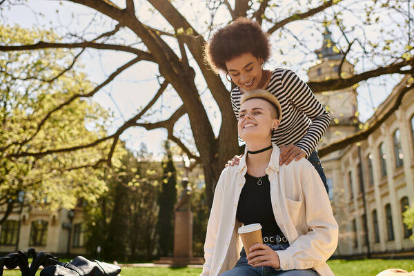 Two young women sitting on grass, casually dressed, sipping coffee.