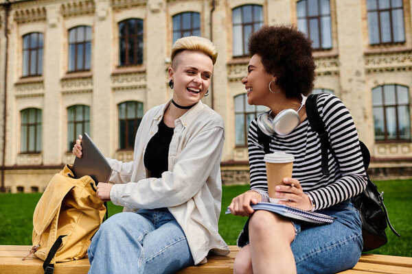 A young multicultural lesbian couple chatting on a bench in a university campus setting.