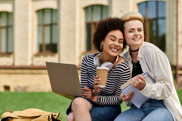 Две молодые женщины, печатающие на ноутбуках, сидя на зеленой траве.