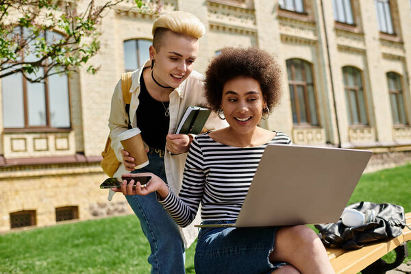 Две женщины, занятые ноутбуком, сидят на скамейке. Их лица показывают внимание и сотрудничество, когда они обсуждают.