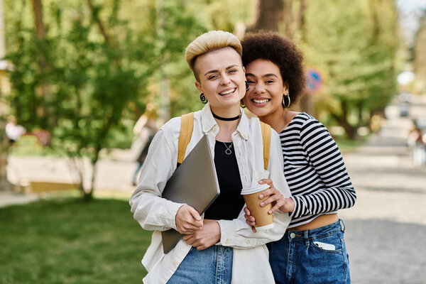Two young women in casual attire stand in a park, holding cups of coffee on a sunny day near a university campus.