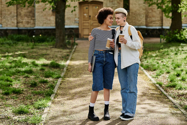 A multicultural lesbian couple in stylish attire walk down a park path, near a university campus, enjoying the outdoor scenery.