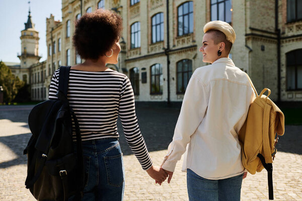 Two young women, in stylish attire, hold hands in front of a building on a university campus.