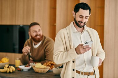Modern bir mutfakta, telefona dalmış, eşiyle modern bir bağ kuran bir adam.