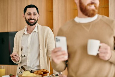 A happy gay couple stands in a kitchen, holding coffee as they spend quality time together in their modern apartment. clipart