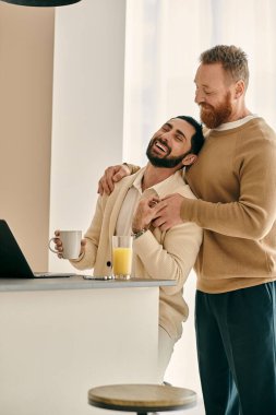 Mutlu bir eşcinsel çift dizüstü bilgisayarın önünde kucaklaşır ve modern bir apartman dairesinde birbirlerine olan sevgilerini gösterirler..