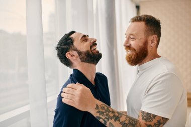 Gündelik giysiler içinde iki mutlu gay pencereli, modern bir oturma odasında gülüyor..