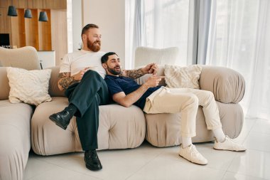 Gündelik giysiler içinde mutlu bir eşcinsel çift modern bir oturma odasındaki koltukta rahatça oturuyor ve birlikte kaliteli vakit geçiriyorlar..