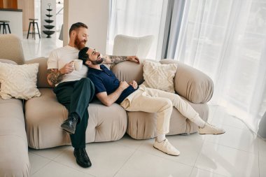 Gündelik giysiler içindeki iki adam, mutlu bir eşcinsel çift, oturma odasındaki modern bir kanepede oturup, bir anlık sevgi ve bağ paylaşıyorlar..