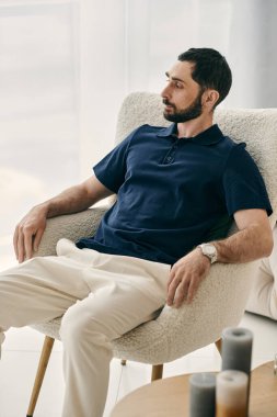 Mavi polo tişörtlü bir adam, bir sandalyede huzur içinde oturur. Modern bir oturma odasında rahatlık ve rahatlığı somutlaştırır..