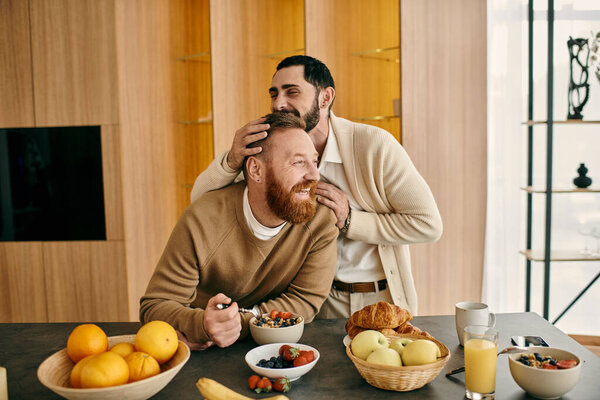 Two men, a happy gay couple, are sitting at a table in a modern apartment, enjoying breakfast together.
