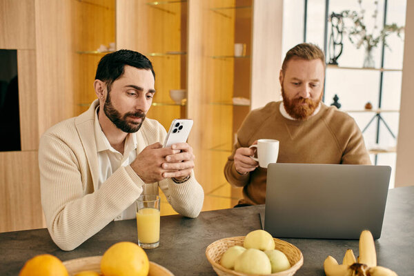 A happy gay couple sits at a table in a modern apartment, engrossed in their phones, enjoying quality time together.
