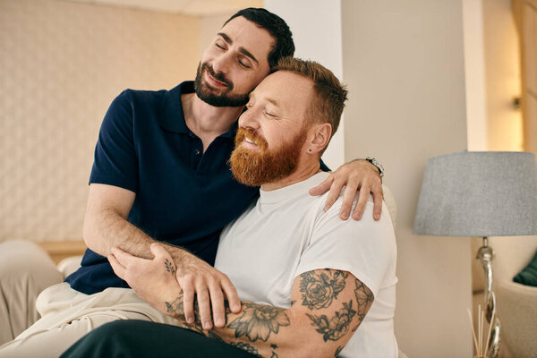 Two men with tattoos embrace warmly on a couch in a modern living room, sharing a moment of love and connection.
