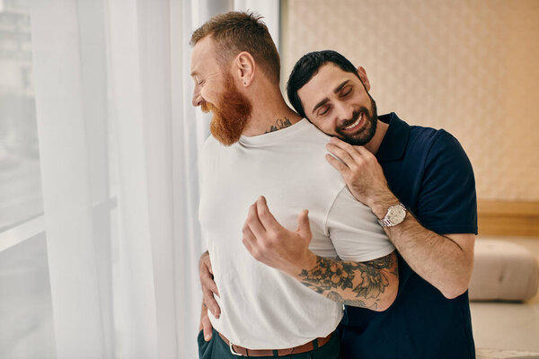 Two men in casual clothes embracing in front of a window, showcasing love and connection in a modern living room.