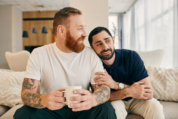 A happy gay couple in casual clothes sitting on a couch, enjoying a cup of coffee in a modern living room.