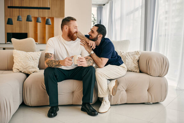 A happy gay couple in casual clothing sit closely together on a couch in a modern living room, enjoying quality time together.