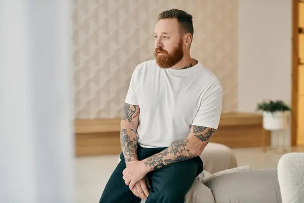 Stock image A man with a beard, sits comfortably on a couch in a modern living room.