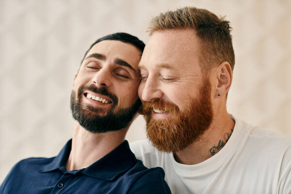 Two men with beards, one in a blue shirt and the other in a striped shirt, are smiling warmly at each other in a cozy living room.