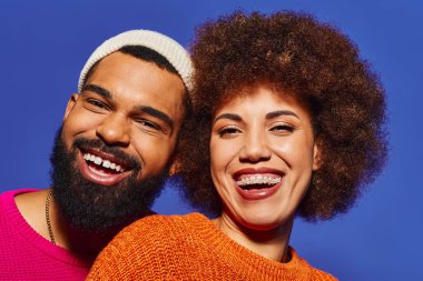Canlı bir şekilde giyinmiş, mavi bir arka planda mutlu bir şekilde gülümseyen Afro-Amerikalı bir çift..
