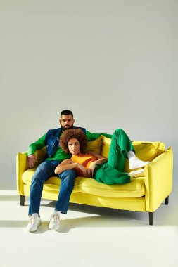 Neşeli bir Afrikalı erkek ve kadın, renkli giyinmiş, sarı bir kanepede gri bir arka plana karşı yan yana otururlar..