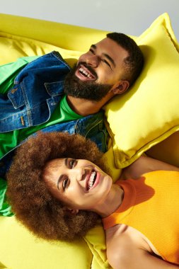 An African American man and woman, dressed in vibrant clothes, are peacefully laying on a yellow pillow against a grey background. clipart