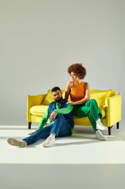 Mutlu bir Afrikalı erkek ve kadın canlı kıyafetler içinde sarı bir kanepede uzanır, arkadaşlıklarını sergilerler..