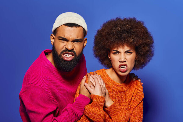 A young African American man and woman, friends in vibrant casual attire, striking a pose together against a blue background.