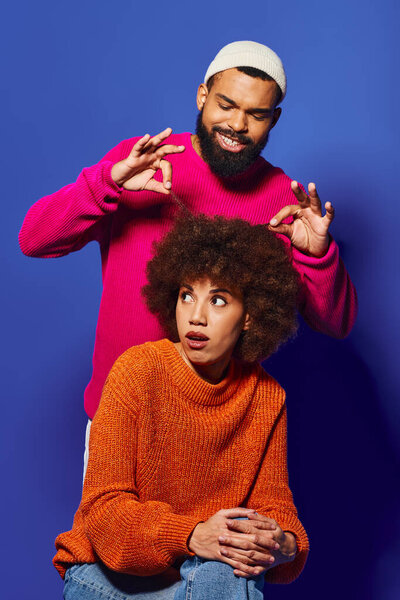 A young African American man and woman, friends, sit closely together in vibrant casual attire against a blue background.